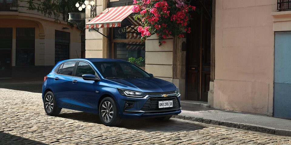 Chevrolet Onix Azul Boreal estacionado em rua de paralelepípedo, com fachada florida ao fundo.