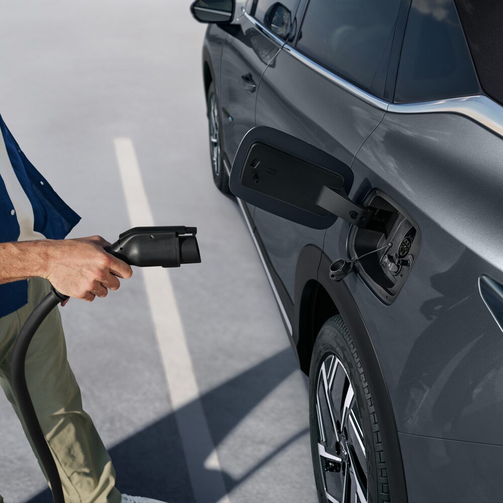 Pessoa conectando o cabo de carregamento em um SUV Chevrolet Captiva EV estacionado, mostrando o detalhe da porta de recarga elétrica.