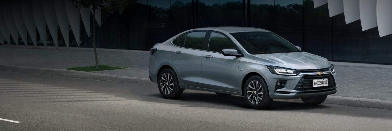 Desan Onix Plus Chevrolet prata estacionado em uma rua, com fachada urbana ao fundo.