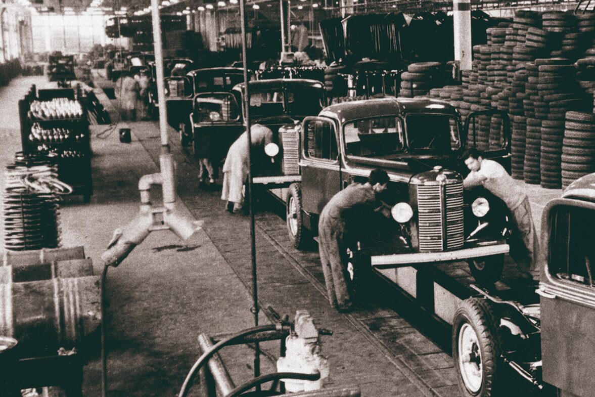 Linha de produção da General Motors do Brasil na década de 1940, com trabalhadores montando automóveis sedã. Os veículos aparecem alinhados na esteira enquanto funcionários ajustam a parte frontal, com pilhas de pneus armazenados ao fundo.