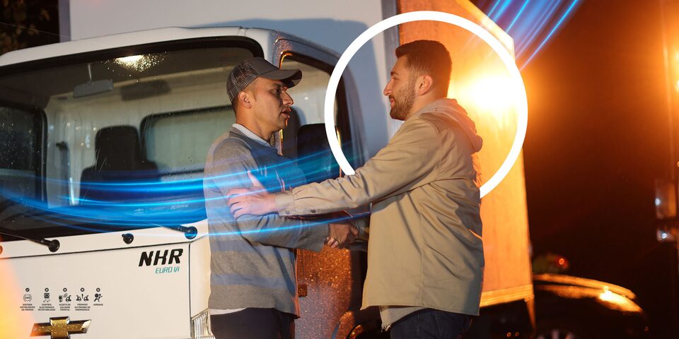 Camión Chevrolet NHR con tecnología OnStar, representado en una escena nocturna donde dos hombres se saludan frente al vehículo.