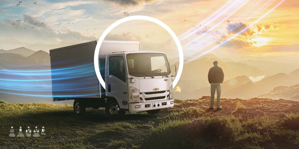 Camión Chevrolet NHR con tecnología OnStar, estacionado en un paisaje montañoso al atardecer junto a un hombre contemplando la vista.