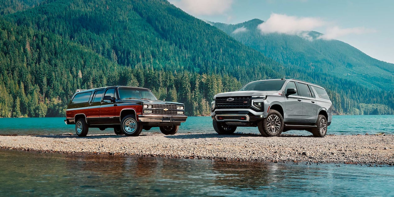 Imagen de una Chevrolet Suburban Vintage al lado de una Chevrolet Suburban Z71, ambas estacionadas a la orilla de un lago a la vera de la montaña.